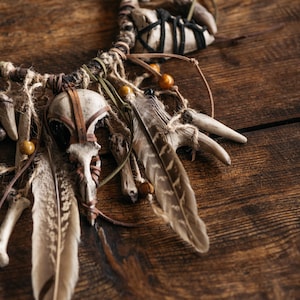 May include: A decorative wall hanging featuring a central bird skull, multiple light-colored animal bones, and brown-patterned feathers. The elements are bound together with twine, leather cords, and small wooden beads, creating a textured arrangement.