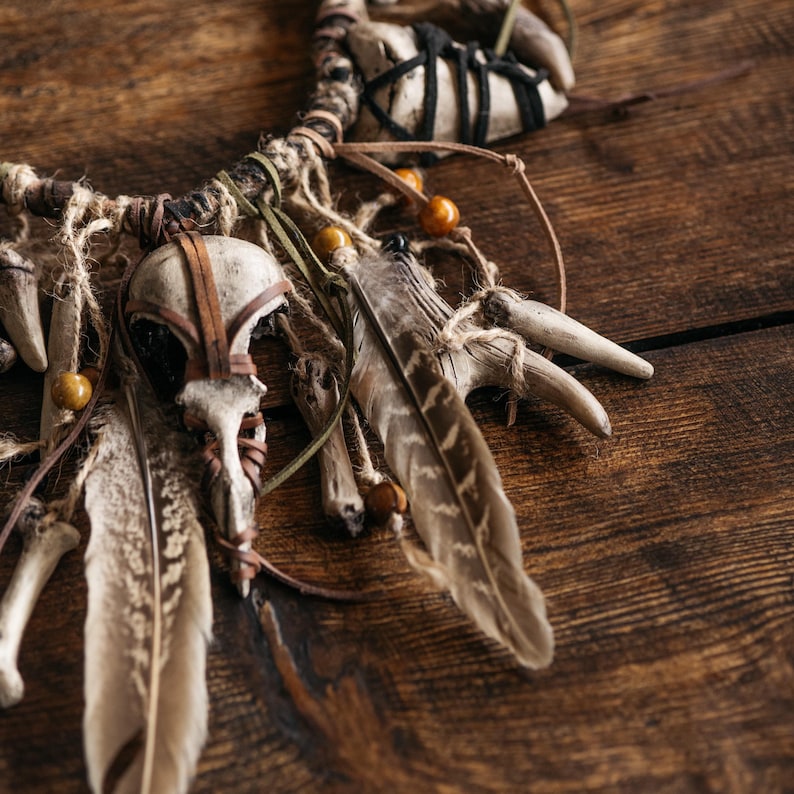 May include: A decorative wall hanging featuring a central bird skull, multiple light-colored animal bones, and brown-patterned feathers. The elements are bound together with twine, leather cords, and small wooden beads, creating a textured arrangement.