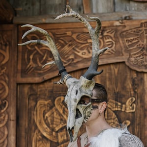 May include: A detailed deer skull mask with large antlers, painted in shades of grey and beige, with rune-like markings. The antlers are attached with twine and a black band. The background features a carved wooden door. The mask is a unique costume piece.