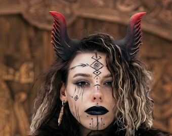 Pode incluir: Uma pessoa com batom preto e pintura facial, usando chifres pretos e vermelhos. Os chifres são pretos com pontas vermelhas. A pessoa está usando uma gola de pele preta. O fundo é uma porta de madeira.