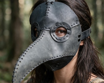Pode incluir: Uma máscara de médico da peste cinzento-escura com um bico longo e pontiagudo, aberturas circulares para os olhos com bordos cosidos e atacadores pretos visíveis ao longo das costuras. A máscara é fixada com uma correia e rebites.