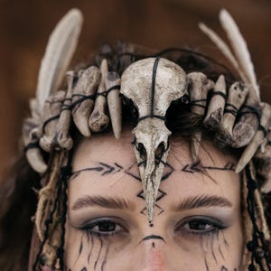 May include: A headpiece constructed from a central bird skull and multiple smaller bone fragments, all secured with black cord. The arrangement features light-colored, weathered bone textures and is designed to be worn across the forehead.