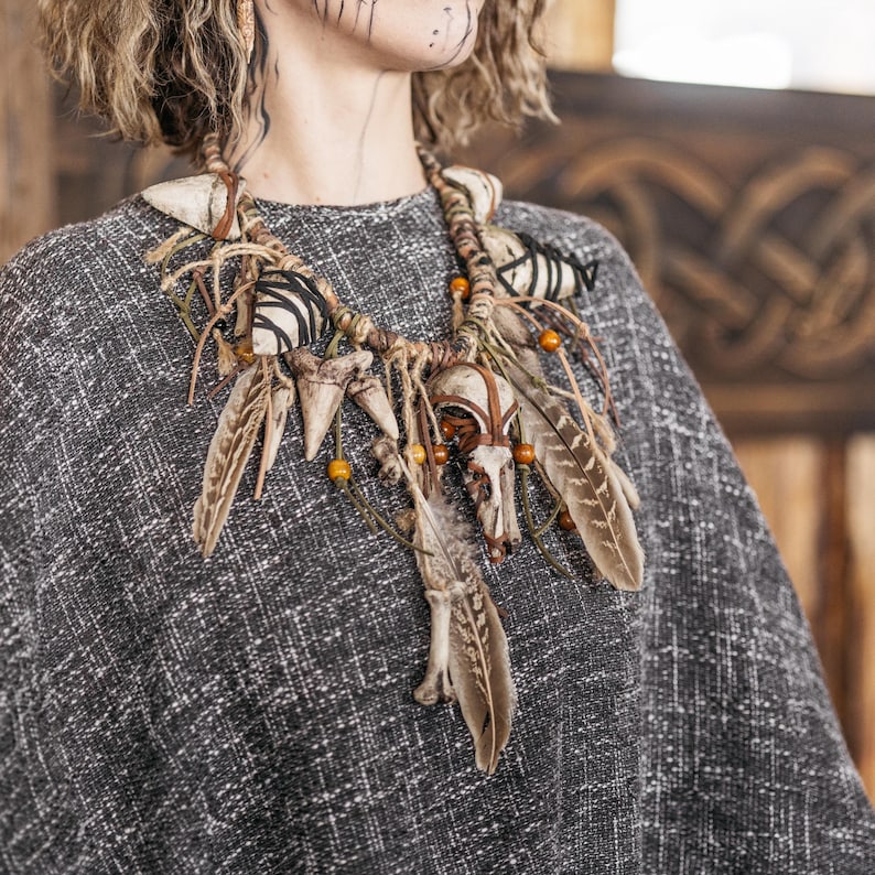 May include: A statement necklace featuring a central animal skull, multiple pointed bone-like elements, and brown patterned feathers. The piece is constructed with wrapped twine, leather cords, and small amber-colored beads, creating a layered design.