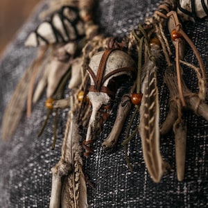 May include: A necklace featuring a central bird skull wrapped in brown leather strips, accented with natural feathers, small wooden beads, and various bone fragments. The components are connected by twine and leather cords, creating a layered design.