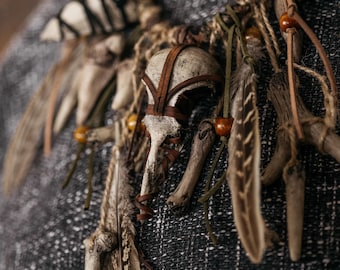 May include: A necklace featuring a central bird skull wrapped in brown leather strips, accented with natural feathers, small wooden beads, and various bone fragments. The components are connected by twine and leather cords, creating a layered design.