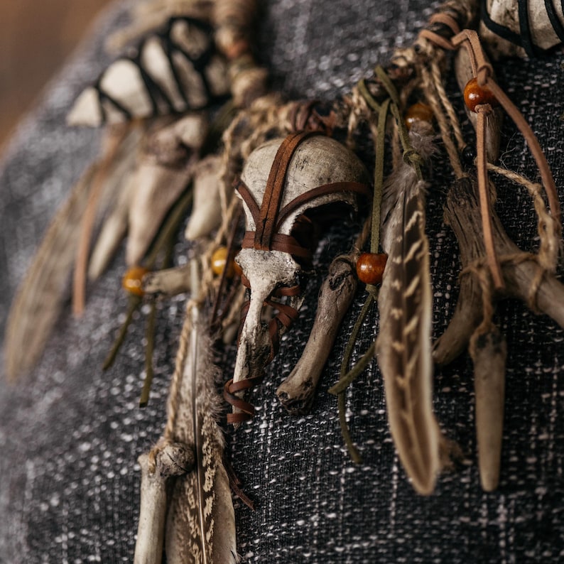 May include: A necklace featuring a central bird skull wrapped in brown leather strips, accented with natural feathers, small wooden beads, and various bone fragments. The components are connected by twine and leather cords, creating a layered design.