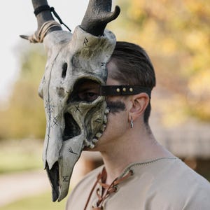 May include: A detailed, handcrafted mask shaped like a skull with attached antlers. The mask is mainly off-white with dark accents and etched symbols. A dark brown leather strap secures the mask. The mask is worn by a person.