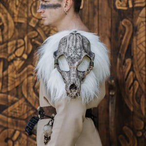 May include: A shoulder mantle featuring a realistic animal skull replica with intricate black runic symbols etched onto the surface. The skull is surrounded by long, thick white faux fur and secured with thin brown cord lacing across the snout.