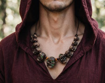 Pode incluir: Um colar com uma série de contas de caveira castanhas esculpidas, intercaladas com pequenas contas de madeira redondas. As caveiras têm detalhes de musgo verde nas órbitas oculares e fendas, enfiadas num fio preto fino à volta do pescoço.