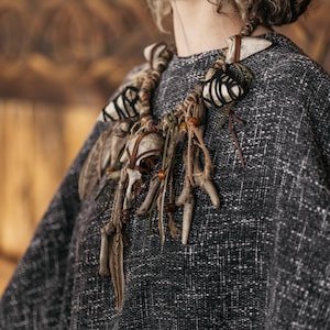 May include: A statement necklace featuring multiple off-white bone-like elements, dark brown leather cords, and small amber-colored beads. The piece is constructed with wrapped twine and organic textures, creating a layered, asymmetrical design.
