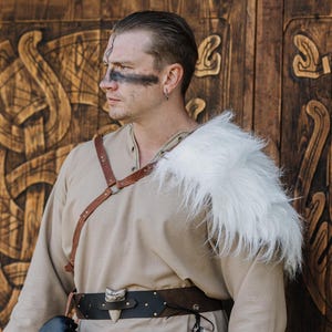 May include: A beige long-sleeved tunic paired with a white faux fur shoulder mantle, a brown leather chest harness, and a dark brown leather belt featuring a prominent white tooth-shaped ornament centered on the front of the waist.