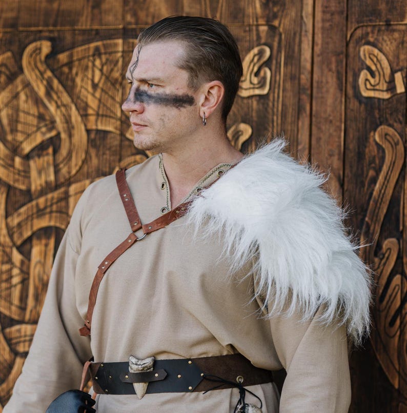 May include: A beige long-sleeved tunic paired with a white faux fur shoulder mantle, a brown leather chest harness, and a dark brown leather belt featuring a prominent white tooth-shaped ornament centered on the front of the waist.