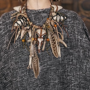 May include: A statement necklace featuring a central animal skull, multiple white teeth, brown feathers, and wooden beads. The components are attached to a thick, braided cord with black thread accents, creating a layered, textured design.