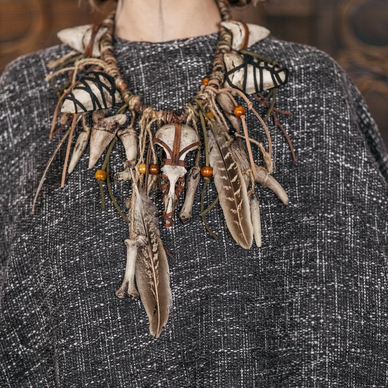 May include: A statement necklace featuring a central animal skull, multiple white teeth, brown feathers, and wooden beads. The components are attached to a thick, braided cord with black thread accents, creating a layered, textured design.