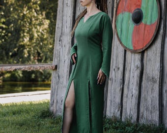 Pode incluir: Um vestido longo verde esmeralda com mangas compridas e decote em V. O vestido apresenta fendas altas em ambos os lados. O vestido é feito de um tecido texturizado e é adequado para reconstituições históricas ou eventos de fantasia.