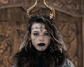 Pode incluir: Uma pessoa com maquiagem escura e chifres dourados. A pessoa está usando uma gola de pele preta e uma jaqueta estampada. O fundo é uma porta de madeira com entalhes. A pessoa tem batom preto e pintura facial tribal.