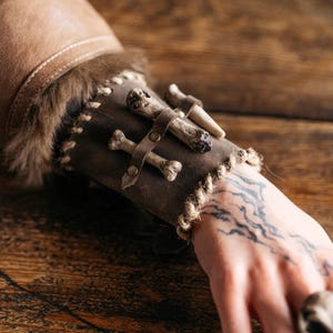 Könnte beinhalten: Ein brauner Lederarmreif mit Knochenakzenten. Der Armreif hat ein genähtes Design mit Fellbesatz und ist mit kleinen knochenförmigen Verzierungen versehen, die mit Lederriemen und Messingnieten befestigt sind. Eine seilartige Verzierung umrandet den Armreif.