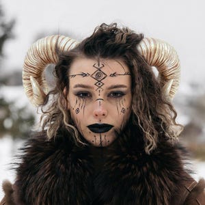 May include: A person with striking face paint and large, curved, golden horns. The person is wearing a brown fur-trimmed garment. Black markings decorate the face, including the lips and around the eyes. The background is blurred, suggesting an outdoor setting.