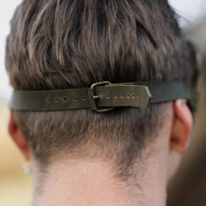 May include: A close-up shot of the back of a person's head, highlighting a dark green leather strap with a metal buckle. The strap, with visible stitching and adjustment holes, is fastened around the head. The background is blurred, drawing attention to the strap's details.