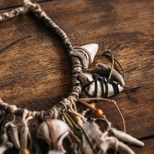 May include: A necklace featuring a braided cord base adorned with multiple off-white tooth-shaped pendants, dark brown and black leather wrappings, thin green and tan cord accents, and small round amber-colored beads integrated into the design.