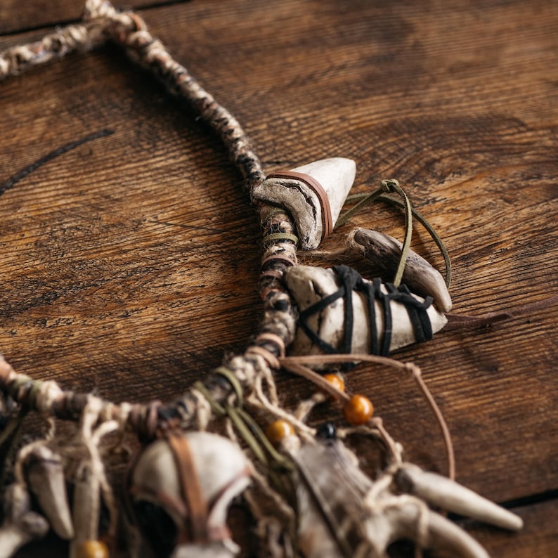 May include: A necklace featuring a braided cord base adorned with multiple off-white tooth-shaped pendants, dark brown and black leather wrappings, thin green and tan cord accents, and small round amber-colored beads integrated into the design.