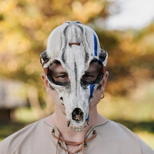 May include: A white and grey skull mask with blue accents and brown leather straps. The mask is designed to resemble an animal skull, with detailed bone structure and eye sockets. The mask is worn by a person wearing a tan shirt.