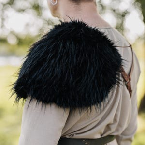 May include: A black faux fur shoulder mantle worn over a light beige long-sleeved garment. The accessory features long, shaggy dark fibers covering the shoulder area, secured with thin brown leather straps visible across the back and side.
