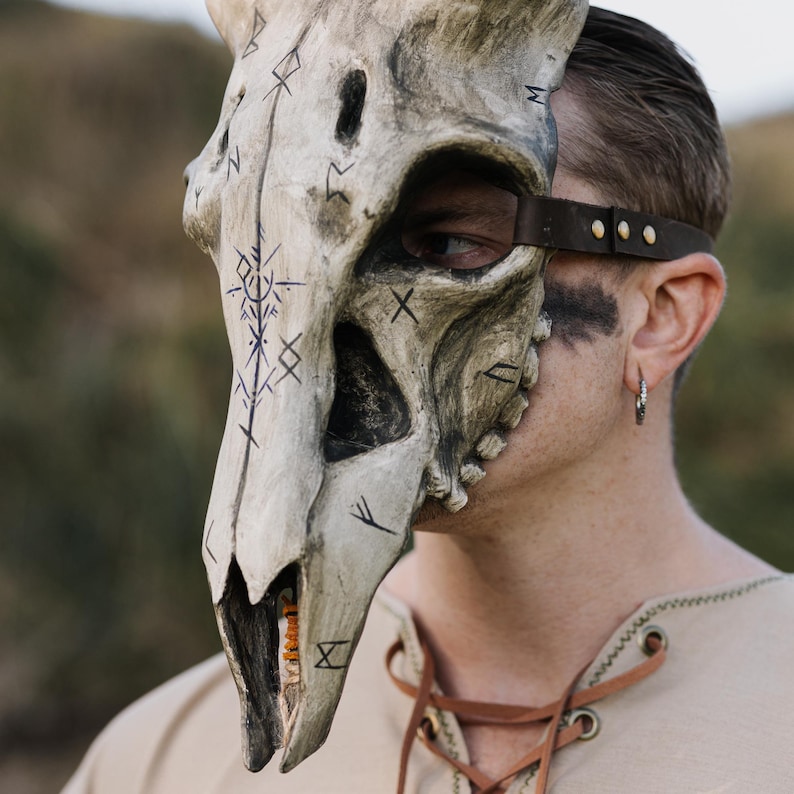 May include: A bone-coloured, half-face mask shaped like a skull, featuring dark markings and a leather strap. The mask has a weathered appearance, suggesting an ancient or fantasy theme. The mask is worn on the face.