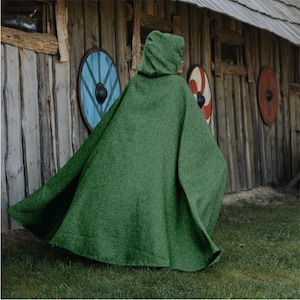May include: A green hooded cloak made of a textured fabric. The cloak is worn outdoors, with a wooden building and decorative shields in the background. The cloak has a wide, flowing design.