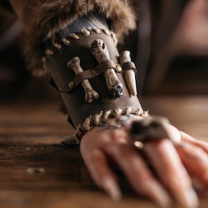 May include: A dark brown leather wrist cuff featuring bone and antler embellishments. The cuff is decorated with small bones and a single antler, held in place by leather straps and metal rivets. A braided rope trim completes the design.