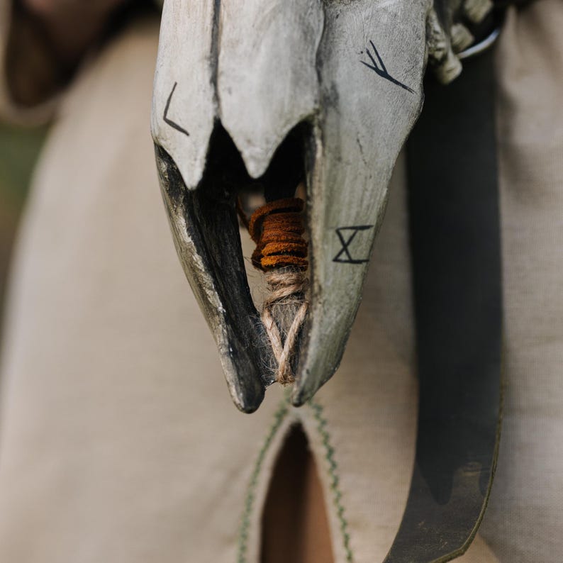 May include: Close-up of a carved, weathered skull with dark markings and a wrapped, orange-brown bundle inside. The skull is attached to a leather strap, suggesting a decorative or ceremonial item.