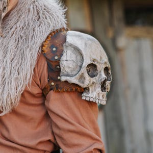 May include: A skull shoulder pauldron, a piece of armor, is attached to a brown garment. The pauldron is made of a skull and leather. The skull is painted white and gray, and the leather is brown with rivets.