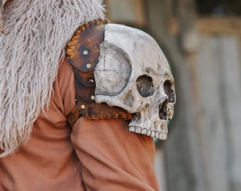 Pode incluir: Uma ombreira de caveira, uma peça de armadura, está presa a uma peça de roupa castanha. A ombreira é feita de uma caveira e couro. A caveira é pintada de branco e cinza, e o couro é castanho com rebites.