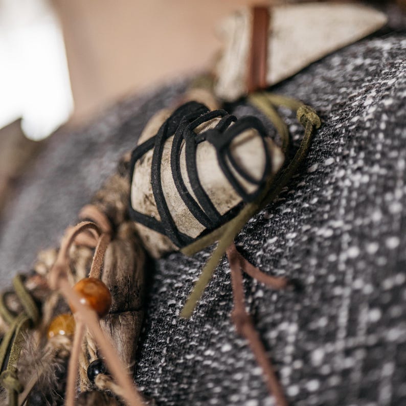 May include: A necklace featuring a central light-colored stone wrapped in black cord, accented with brown leather strips, olive green cord, small amber-colored beads, and dark wooden elements, all suspended on a textured gray fabric surface.