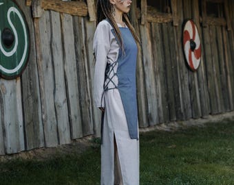 Pode incluir: Um vestido de linho de cor clara com uma sobreposição azul estilo avental, com cadarços nas laterais. O vestido tem mangas compridas e uma fenda alta. O fundo apresenta uma estrutura de madeira e escudos decorativos, sugerindo um tema histórico ou viking.
