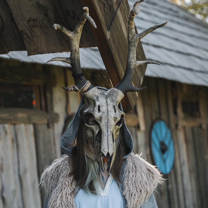 May include: A detailed deer skull mask with large antlers. The mask is painted in shades of grey and beige, with dark markings. The person is wearing a grey hooded cloak with a fur collar, set against a rustic wooden building.