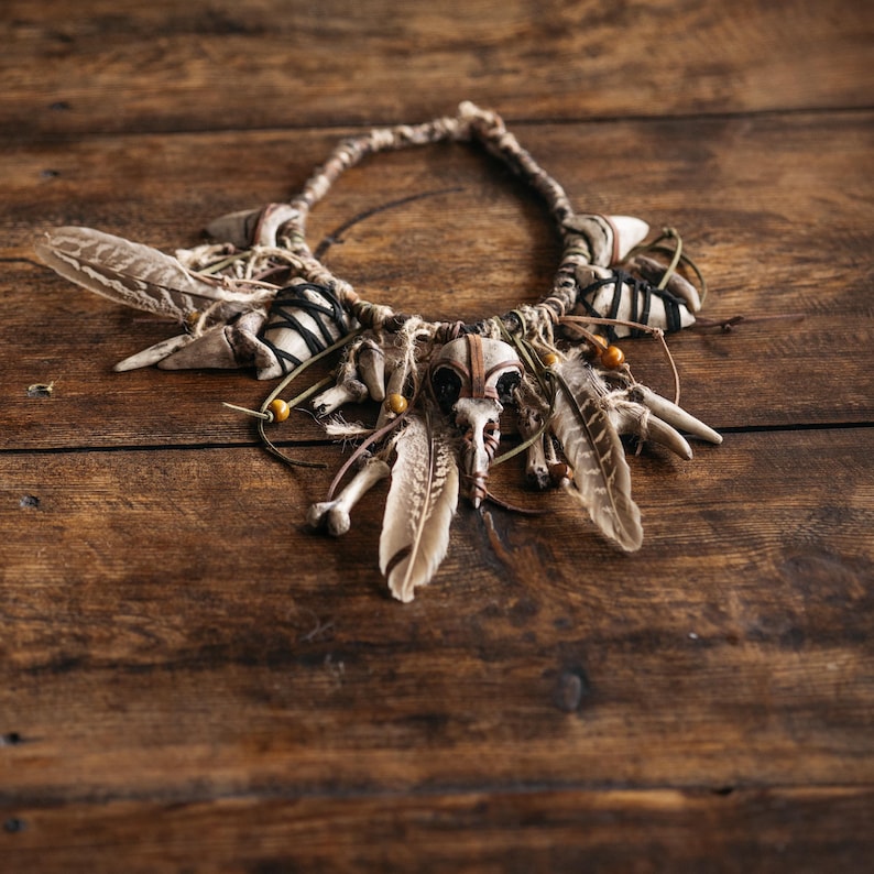May include: A necklace featuring a central bird skull, natural feathers, and small bones. The piece is constructed with tan cord, black thread wrappings, and small wooden beads, creating a layered arrangement of organic elements and textures.