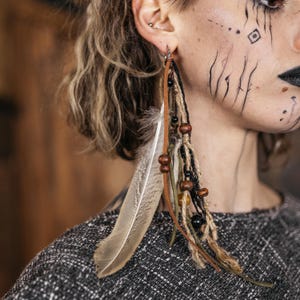 May include: Long, dangling earrings with a bohemian style. The earrings feature a large, light-colored feather, wooden beads, and strands of brown, black, and green materials. The earrings are attached to a silver-colored hook.