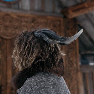 Könnte beinhalten: Ein Kostüm mit großen, gebogenen schwarzen Hörnern, einem Pelzkragen und lockigem braunem und blondem Haar. Die Person trägt eine graue Tweedjacke. Der Hintergrund umfasst eine Holztür mit geschnitzten Details.