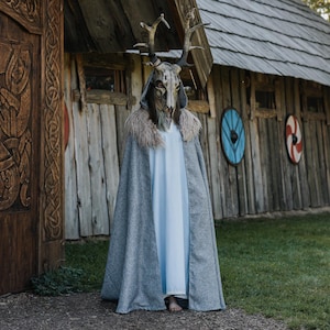 May include: A person wearing a grey cloak with a faux fur collar and a deer skull mask with antlers. The person is standing in front of a wooden building with decorative shields.