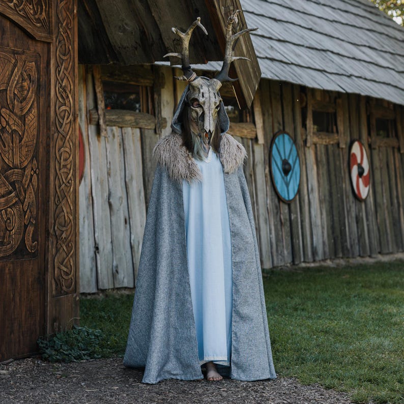 May include: A person wearing a grey cloak with a faux fur collar and a deer skull mask with antlers. The person is standing in front of a wooden building with decorative shields.