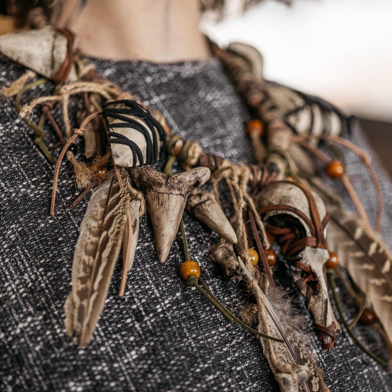 May include: A statement necklace featuring natural elements including pointed bone-like structures, brown feathers, and small amber-colored beads. The components are bound together with brown leather cords and tan twine, with black thread wrapped around some pieces.