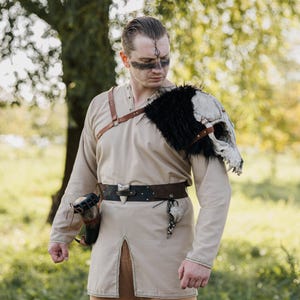 May include: A beige long-sleeved tunic with a front slit, worn with a brown leather harness and a dark brown belt. A large animal skull with black faux fur is attached to the left shoulder. A horn-shaped container hangs from the belt on the right.
