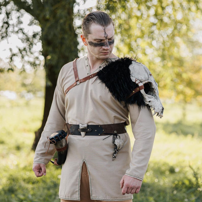 May include: A beige long-sleeved tunic with a front slit, worn with a brown leather harness and a dark brown belt. A large animal skull with black faux fur is attached to the left shoulder. A horn-shaped container hangs from the belt on the right.