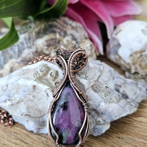 Könnte beinhalten: Eine handgefertigte Kupferdraht-Anhänger-Halskette mit einem tropfenförmigen, polierten, lila Edelstein. Der Edelstein ist in aufwendiger Kupferdrahtweberei eingefasst und hängt an einer zarten Kupferkette. Der Anhänger wird auf einer Steinoberfläche präsentiert.