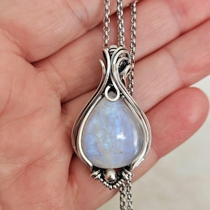 Op de afbeelding: Een druppelvormige maansteen hanger omvat door ingewikkeld zilveren draadwerk. De hanger bevat een gladde, iriserende steen met blauwe flitsen en hangt aan een zilveren ketting. Het metaalwerk bevat decoratieve krullen en een kleine zilveren kraal aan de onderkant.