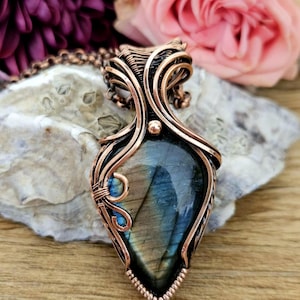 May include: A teardrop-shaped labradorite gemstone pendant encased in intricate copper wire wrapping. The stone displays iridescent blue flashes against a dark grey base. The pendant hangs from a matching copper chain, featuring detailed metalwork.