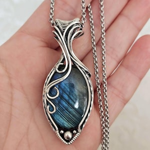 Op de afbeelding: Een hangerketting met een ovale labradoriet edelsteen met blauwe en grijze iridiscentie, gevat in ingewikkeld zilveren draadwerk. De hanger hangt aan een zilveren ketting en toont gedetailleerd metaalwerk en een kleine zilveren kraal aan de basis.