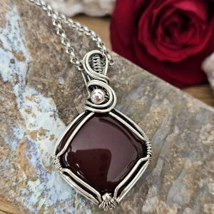 Può includere: Una collana con pendente caratterizzata da una gemma liscia di forma quadrata color marrone scuro, racchiusa in una montatura in filo d'argento. Il pendente è appeso a una catena d'argento, con fili che formano complessi intrecci.