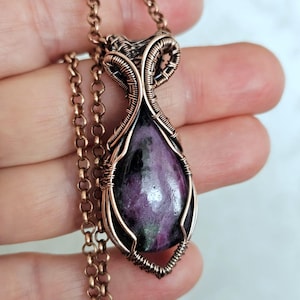 Könnte beinhalten: Eine handgefertigte Halskette mit Anhänger, umwickelt mit Kupferdraht. Der Anhänger zeigt einen tropfenförmigen, lila und grünen Edelstein. Die Halskette hat eine passende kupferfarbene Kette, die ein künstlerisches Schmuckstück bildet.